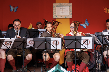 A brass band performs in front of a red backdrop decorated with butterflies, featuring musicians in traditional attire playing various instruments.