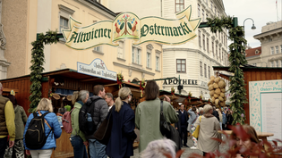 People gather at a bustling outdoor Easter market with festive decorations and vendor stalls in a European street setting.
