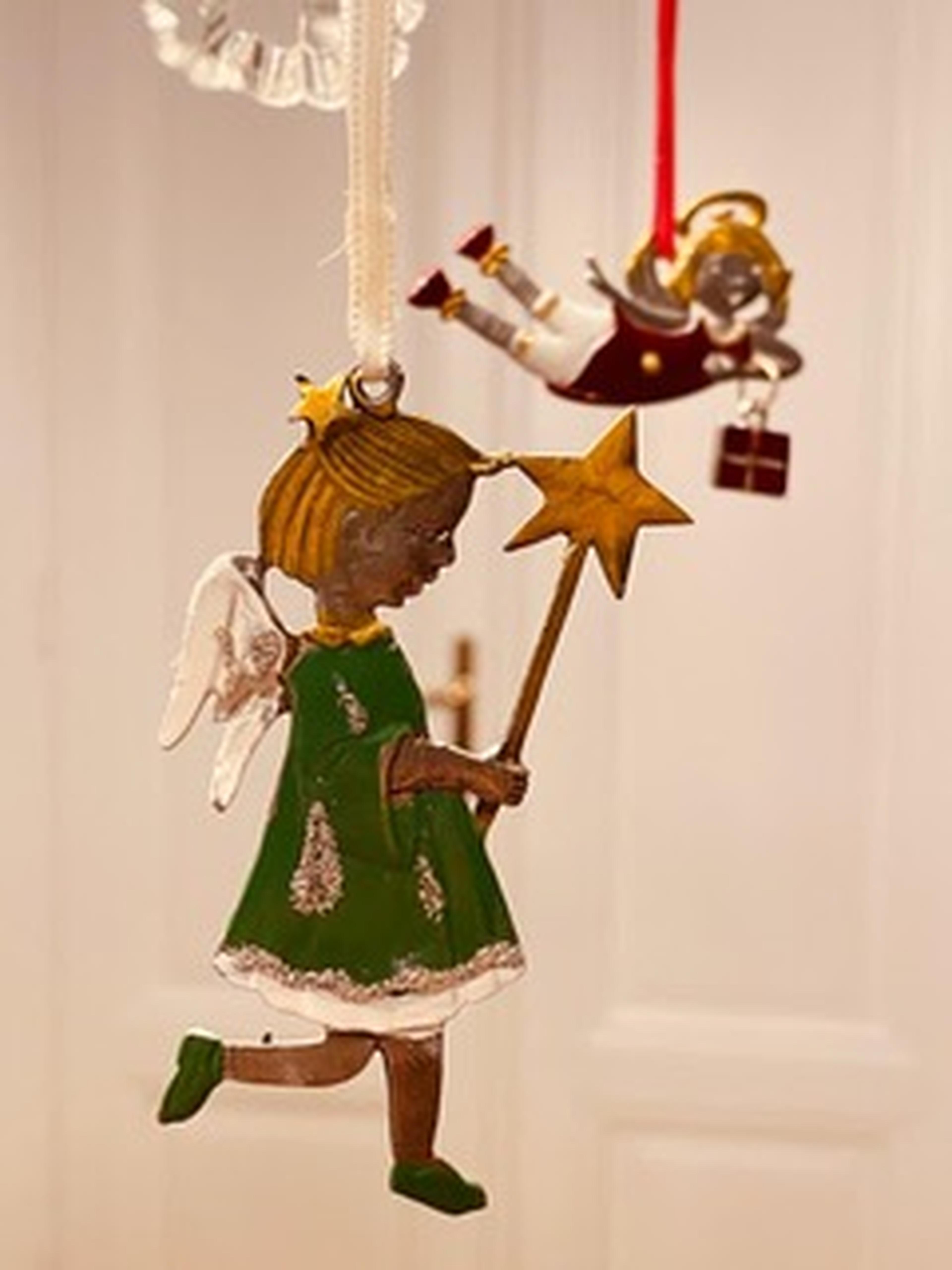 Ornate angel ornaments hanging; one in green with a star wand, another in red holding a gift, against a blurred background.