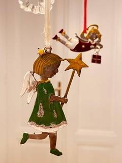 Ornate angel ornaments hanging; one in green with a star wand, another in red holding a gift, against a blurred background.