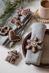 Gray napkins with brown felt holiday ornaments shaped like gingerbread figures and snowflakes, next to a cup on a light table.