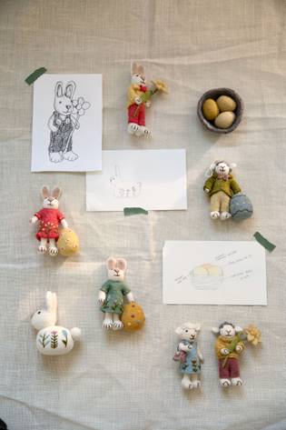 Handmade rabbit dolls in colorful outfits arranged on fabric, with sketches and a basket of eggs.
