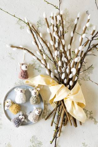 A bundle of pussy willow branches tied with a yellow ribbon, next to a plate of small, colorful, knitted animal ornaments on a textured surface.