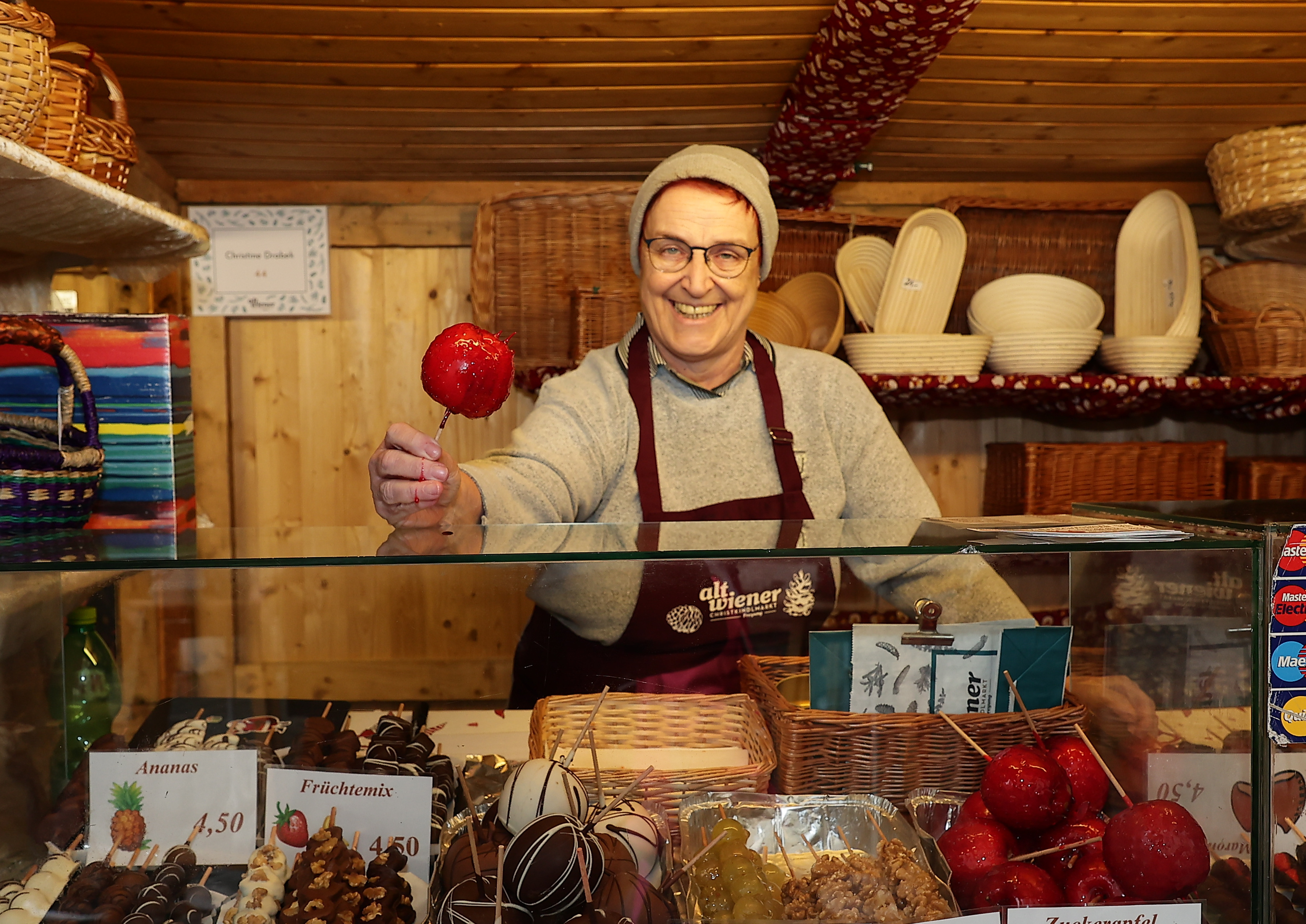 Smiling vendor in a cozy wooden booth holds a red candy apple. Baskets and confections are displayed around, creating a festive atmosphere.