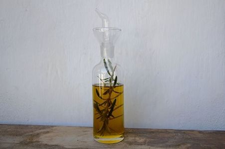 Glass bottle with spout containing olive oil infused with rosemary sprigs, placed on a wooden surface against a plain white wall.