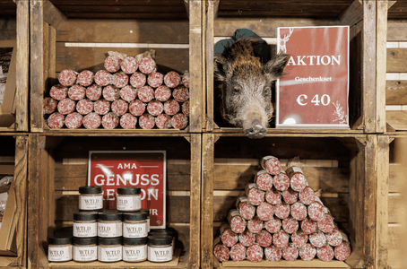 Rustic wooden shelves with stacked salami, jars, and a wild boar head beside a red "Aktion" gift set sign reading €40.