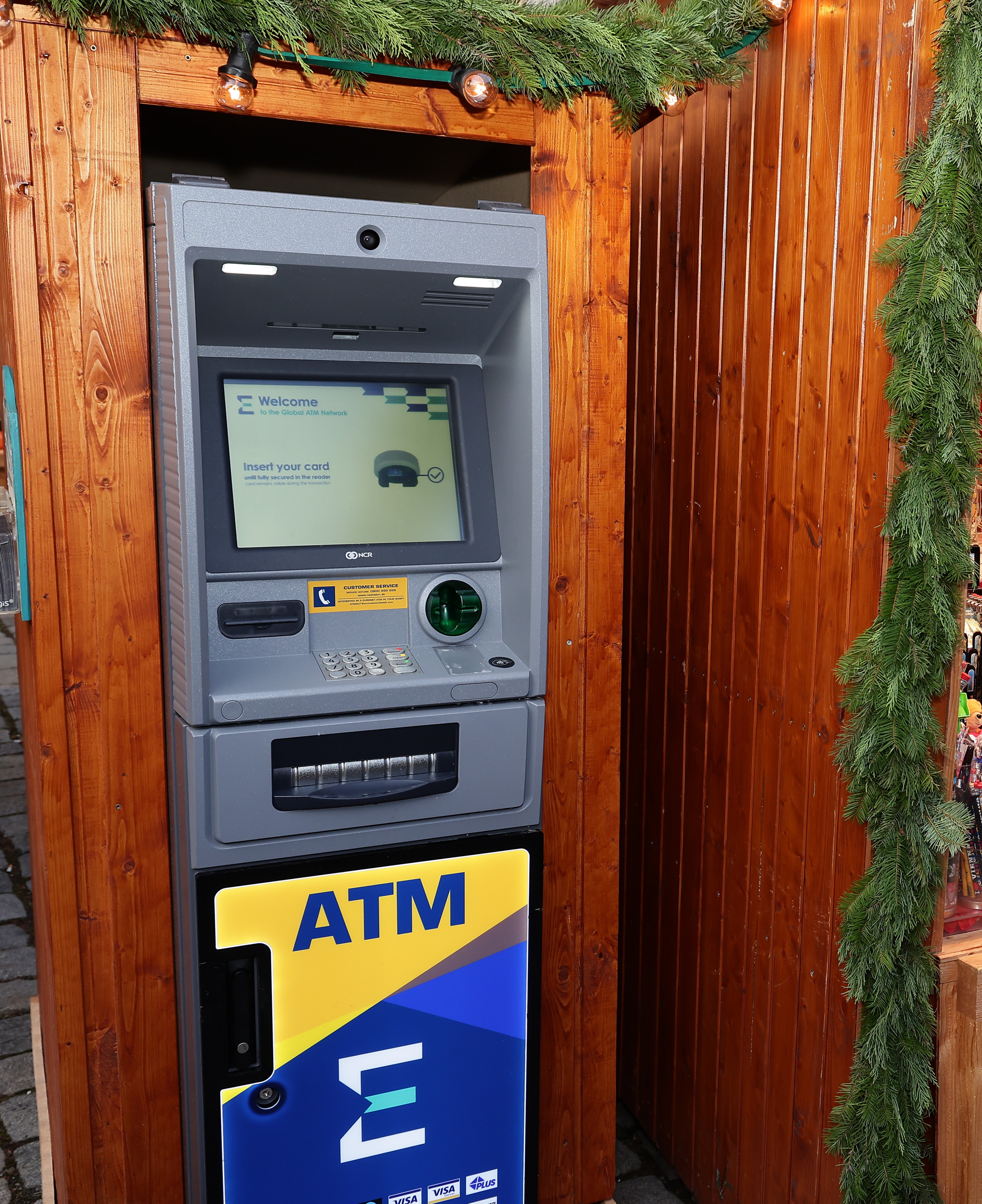 ATM machine in a wooden booth with a screen displaying "Insert your card." Greenery decorates the top of the booth.