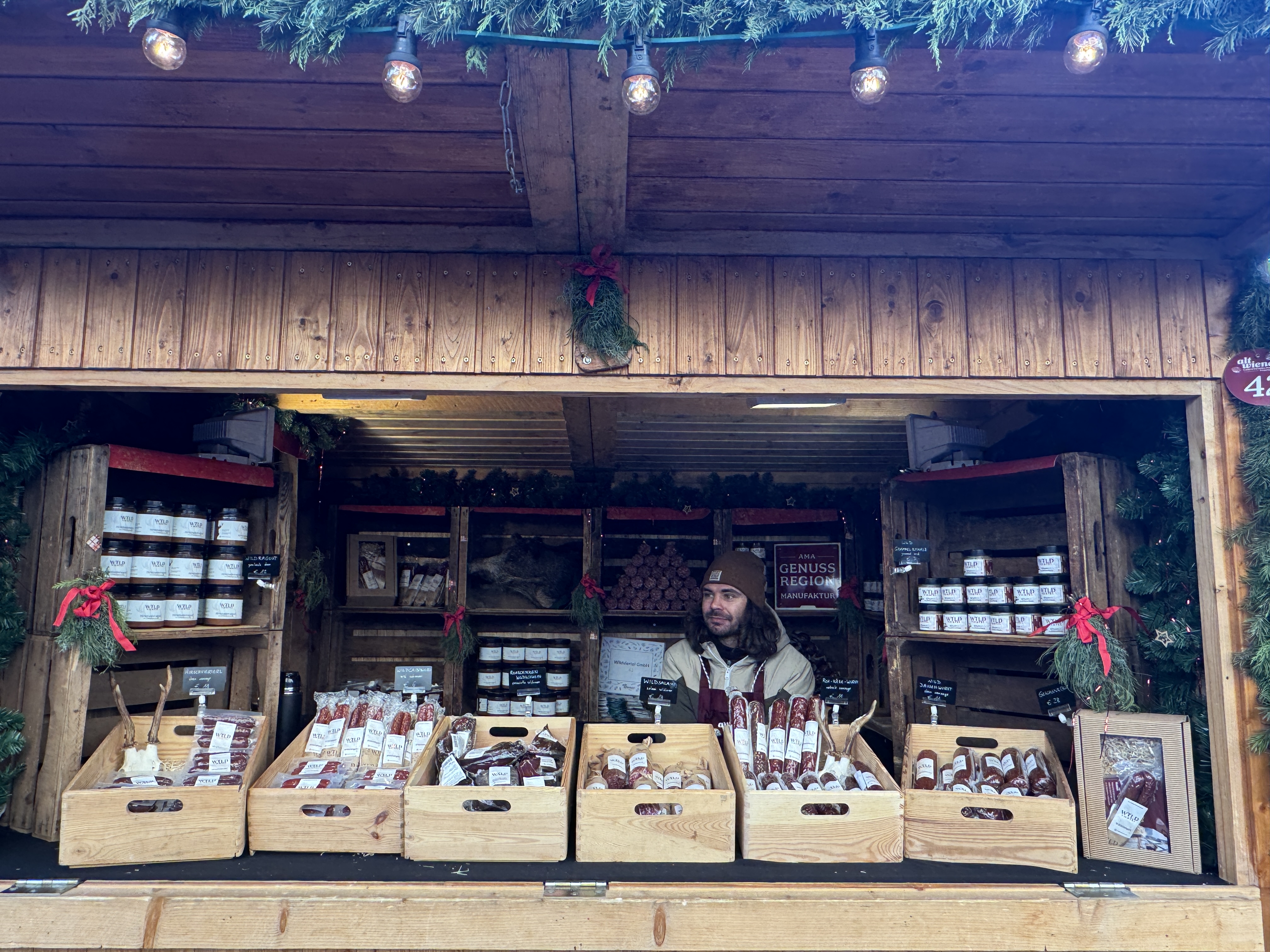 A vendor in a wooden market stall sells various sausages and condiments, with festive decorations and lights overhead.