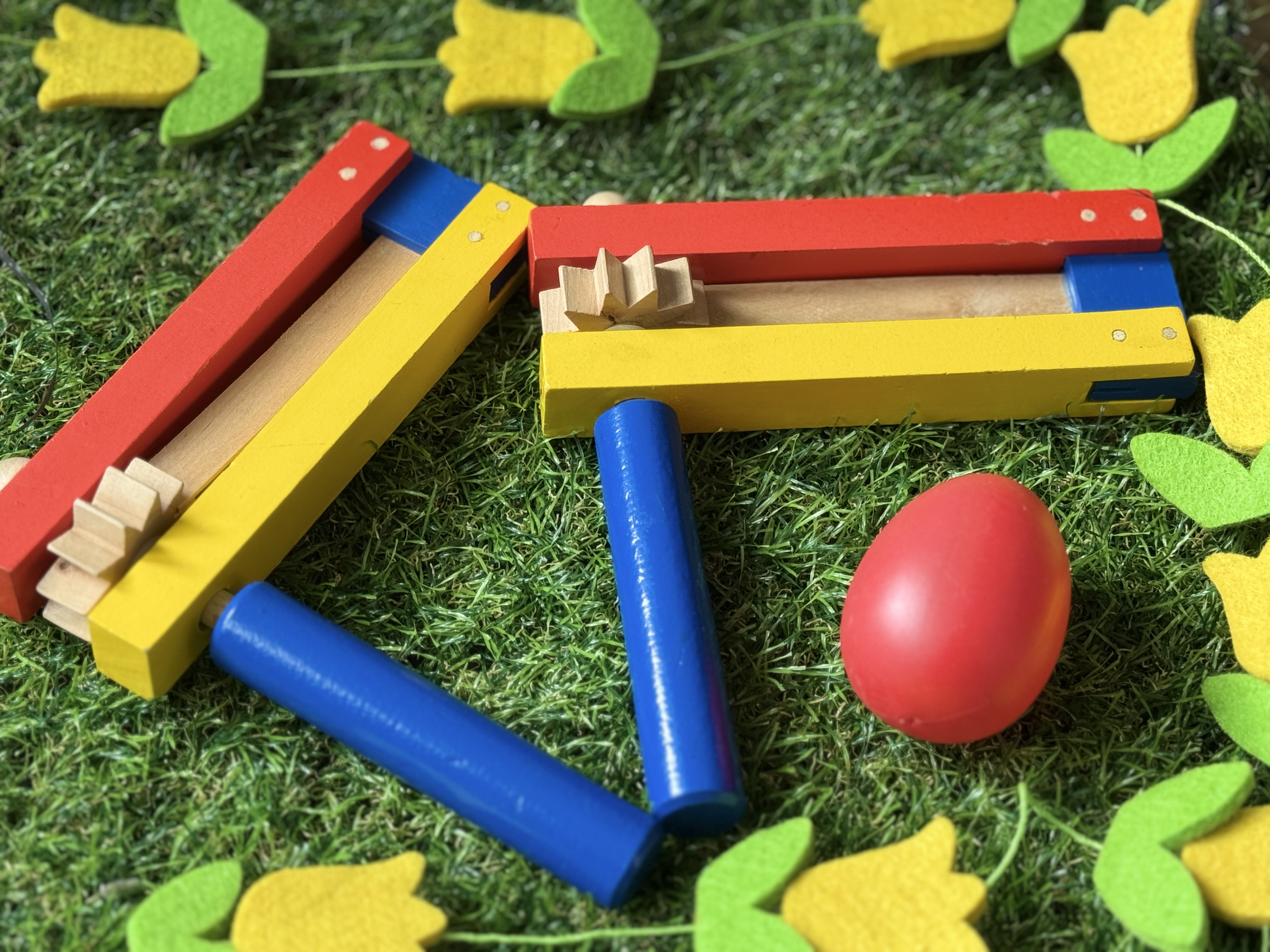 Colorful wooden noisemakers and a red egg sit on grass, surrounded by yellow felt tulips, creating a festive and playful scene.