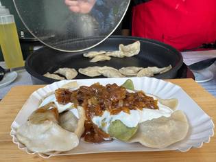 A plate of mixed dumplings topped with sour cream and caramelized onions, with more dumplings cooking in a pan in the background.