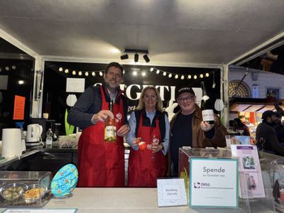 Three people in red aprons stand at a booth, smiling. They hold a bottle, glass, and jar. Donation box visible on the counter.
