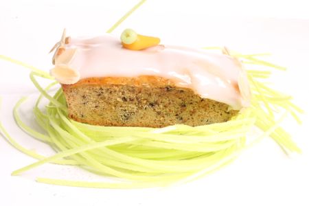 Carrot cake slice with white icing, topped with almond flakes and a carrot decoration, surrounded by green edible grass on a white background.