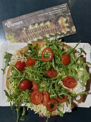 A loaded open-faced langos with cherry tomatoes, arugula, and cheese on a paper plate. A Vienna Christmas market flier is in the background.