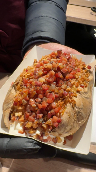 A loaded baked potato in a tray, topped with crispy diced bacon and a dollop of sour cream, held by a person in a dark sleeve, evoking warmth and comfort.