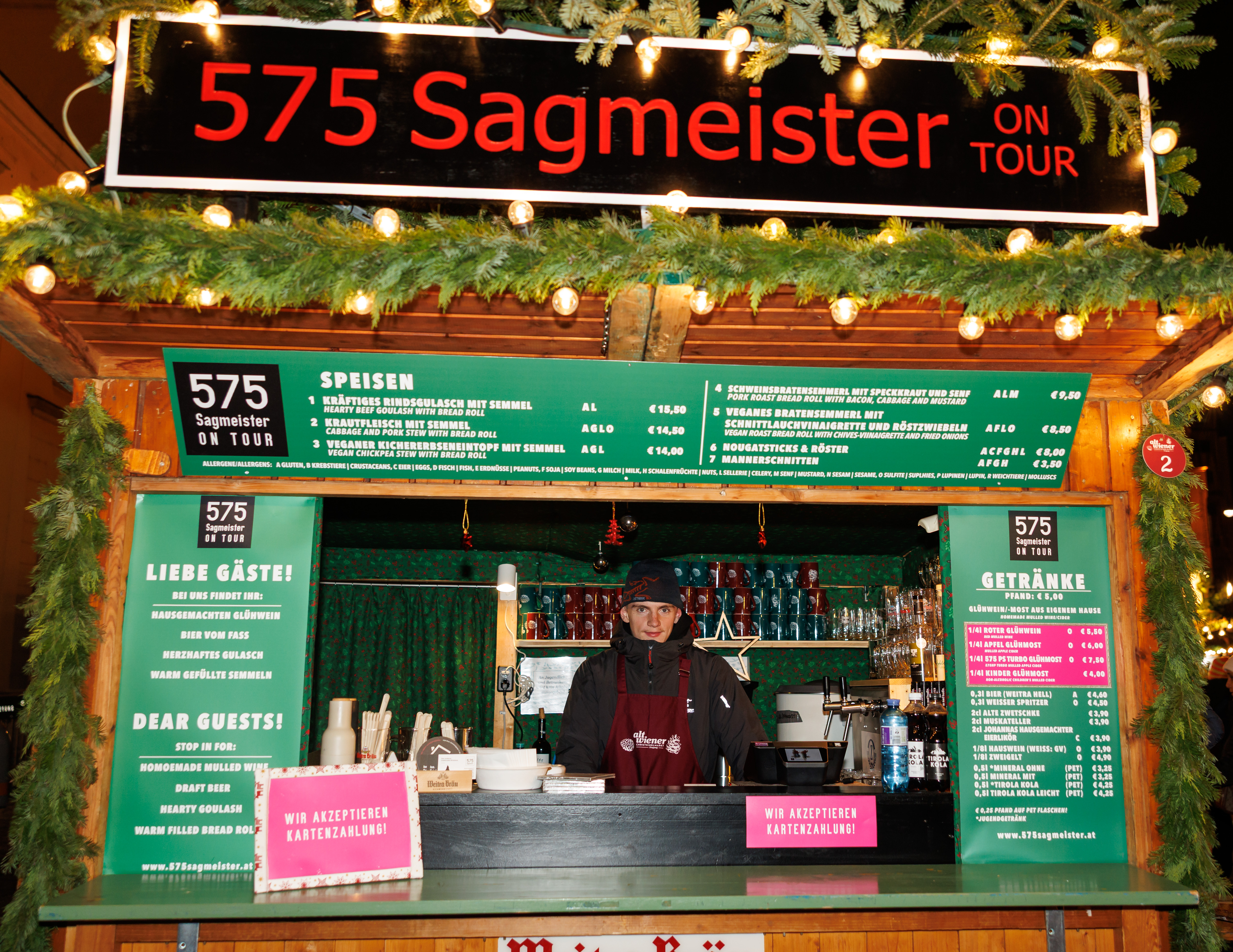 A festive Christmas market food stall labeled “575 Sagmeister On Tour” shows a person standing behind the counter surrounded by menus, greenery, and warm string lights.