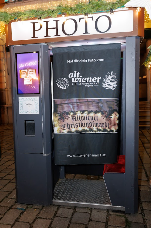 Photo booth at an outdoor Christmas market, decorated with green garlands. A screen displays "Say Cheese!" while a curtain reads "Alt Wiener Christkindlmarkt."