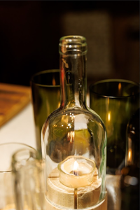 A lit candle sits inside an empty glass bottle surrounded by dark green glasses on a table. The warm, soft glow creates a cozy and intimate atmosphere.