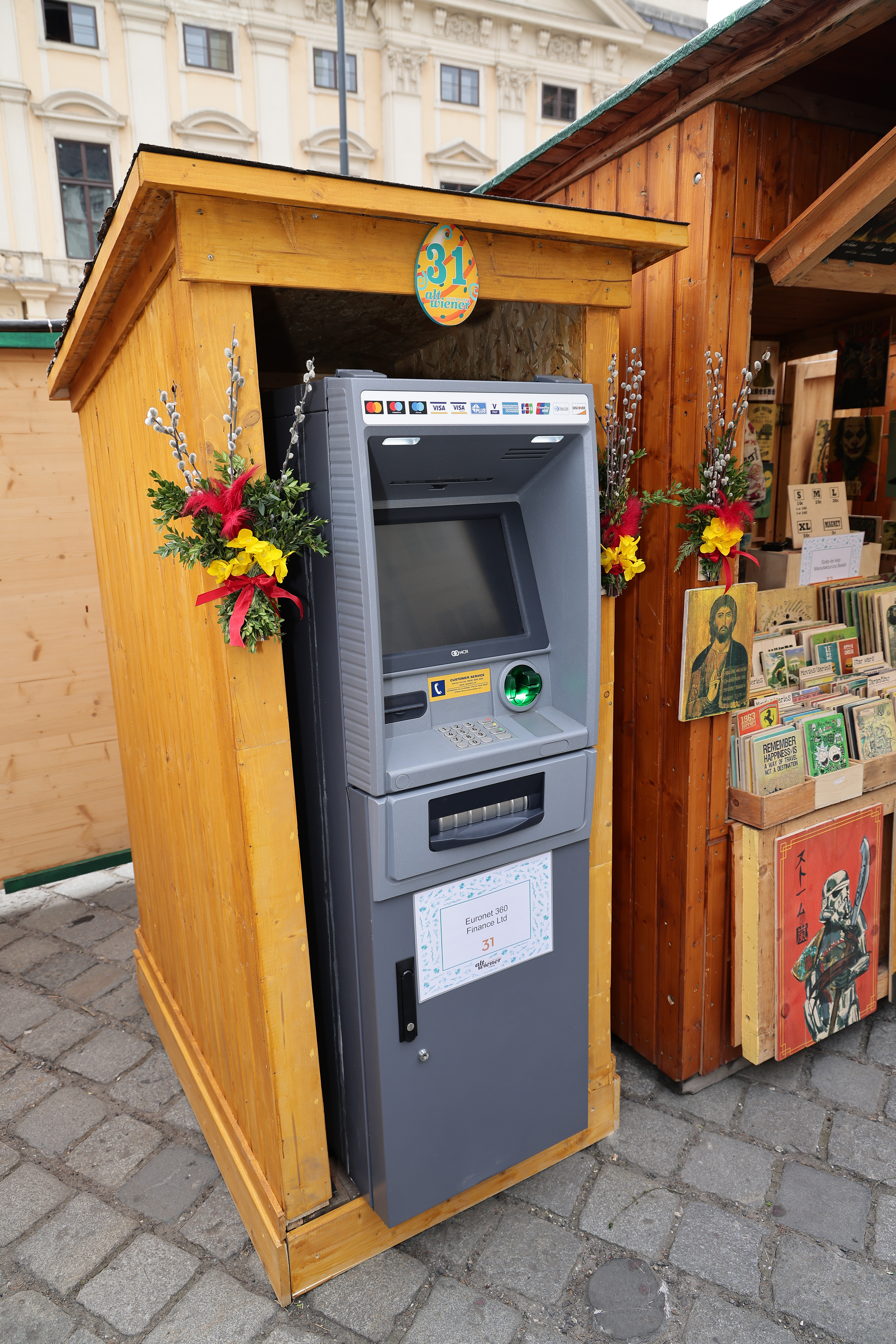 ATM inside a wooden booth, decorated with flowers, on a cobblestone street with a building facade in the background.