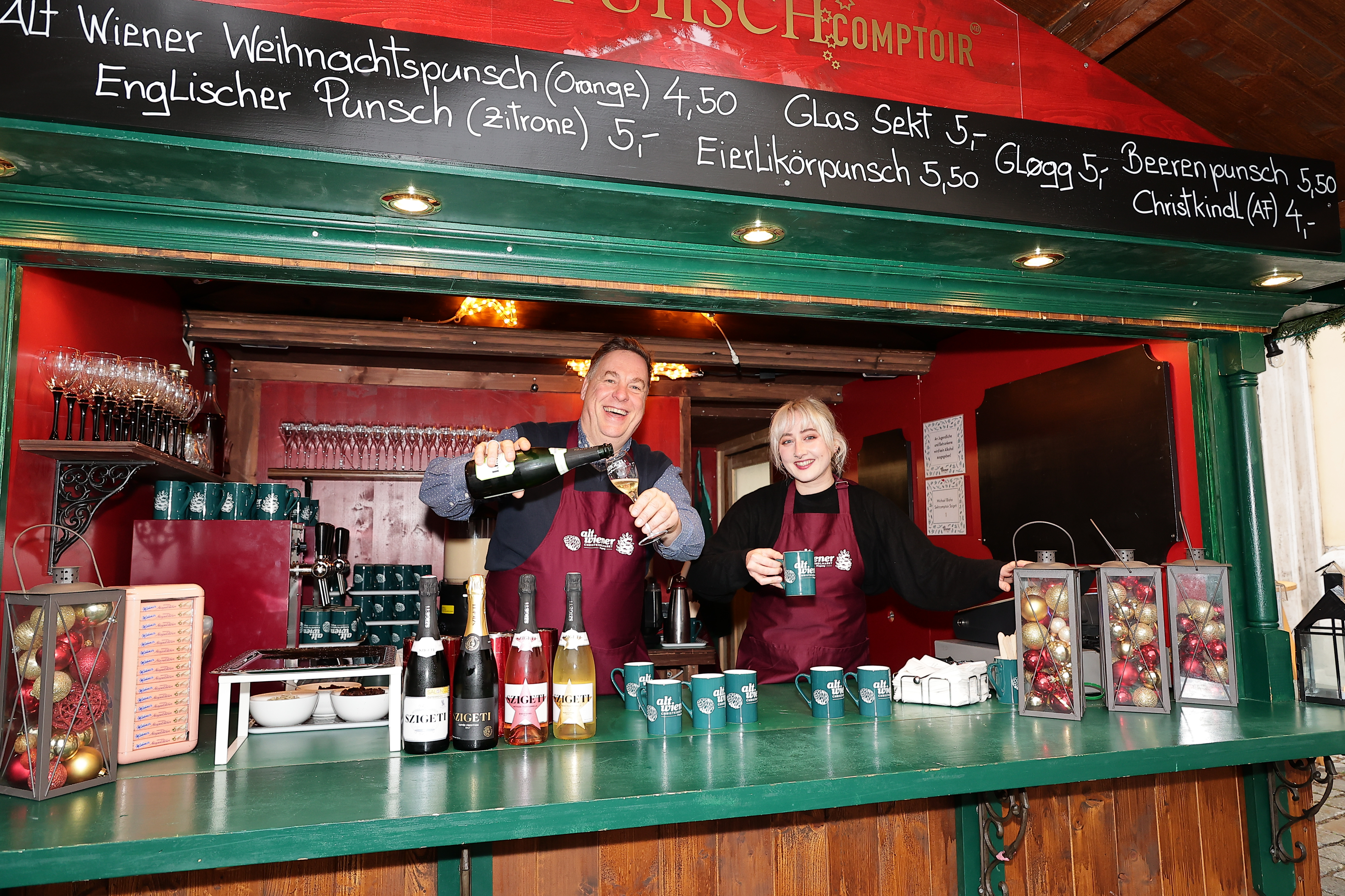 Two people in aprons serve drinks at a festive market stall decorated with Christmas ornaments and price signs.