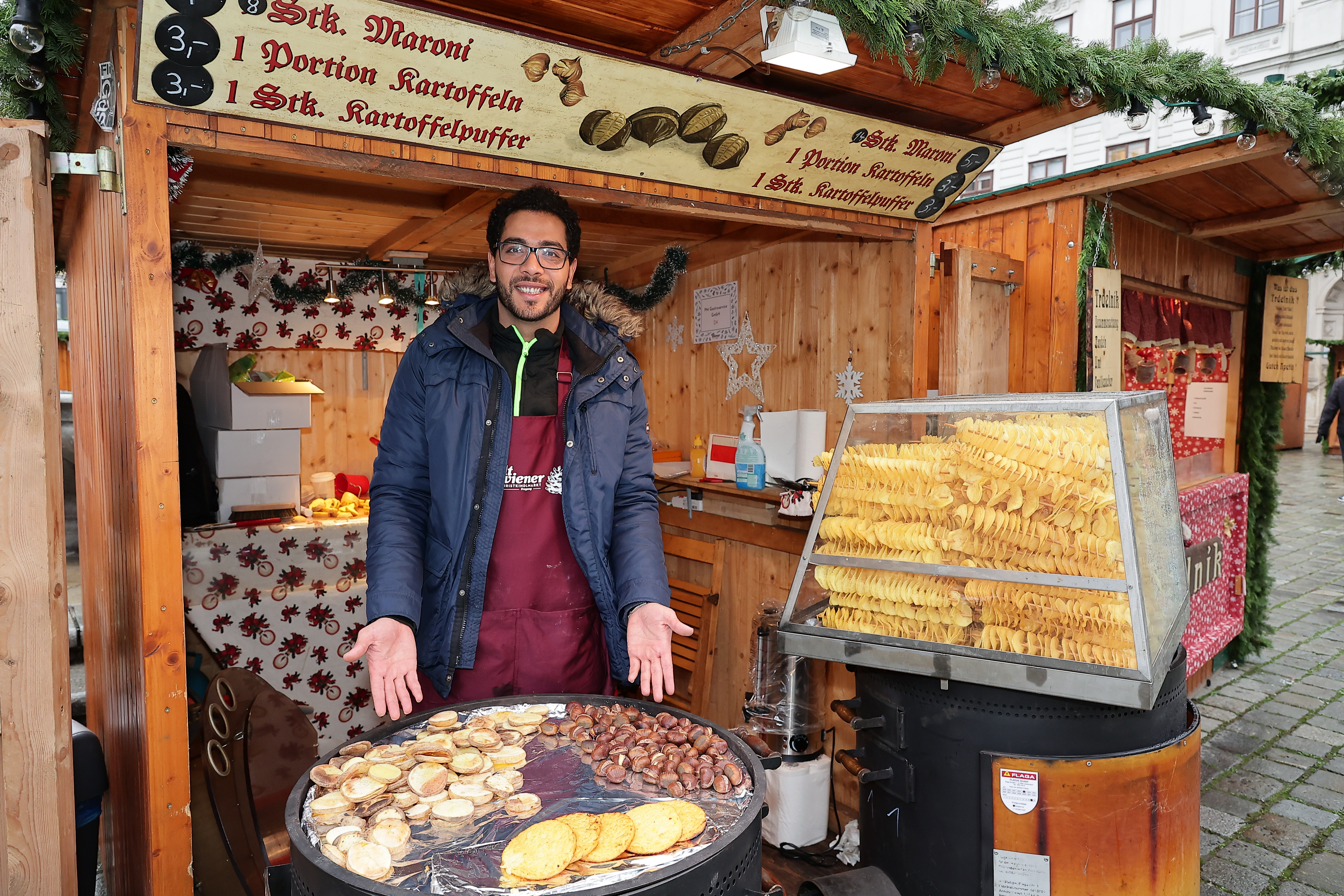 A vendor at a festive market stall sells roasted chestnuts, potato pancakes, and spiral potatoes on a rainy day.
