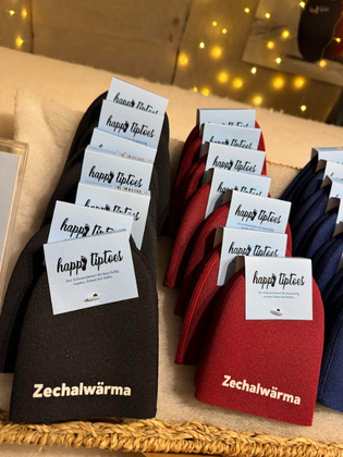 Black, red, and blue "Zechalwärma" slippers with "happy tiptoes" tags are neatly arranged in rows against a warm, softly lit background.