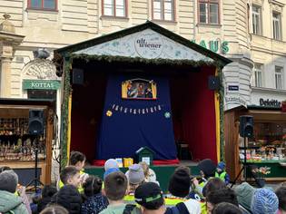 Children watch a puppet show on an outdoor stage decorated with greenery, set against historic buildings and market stalls.