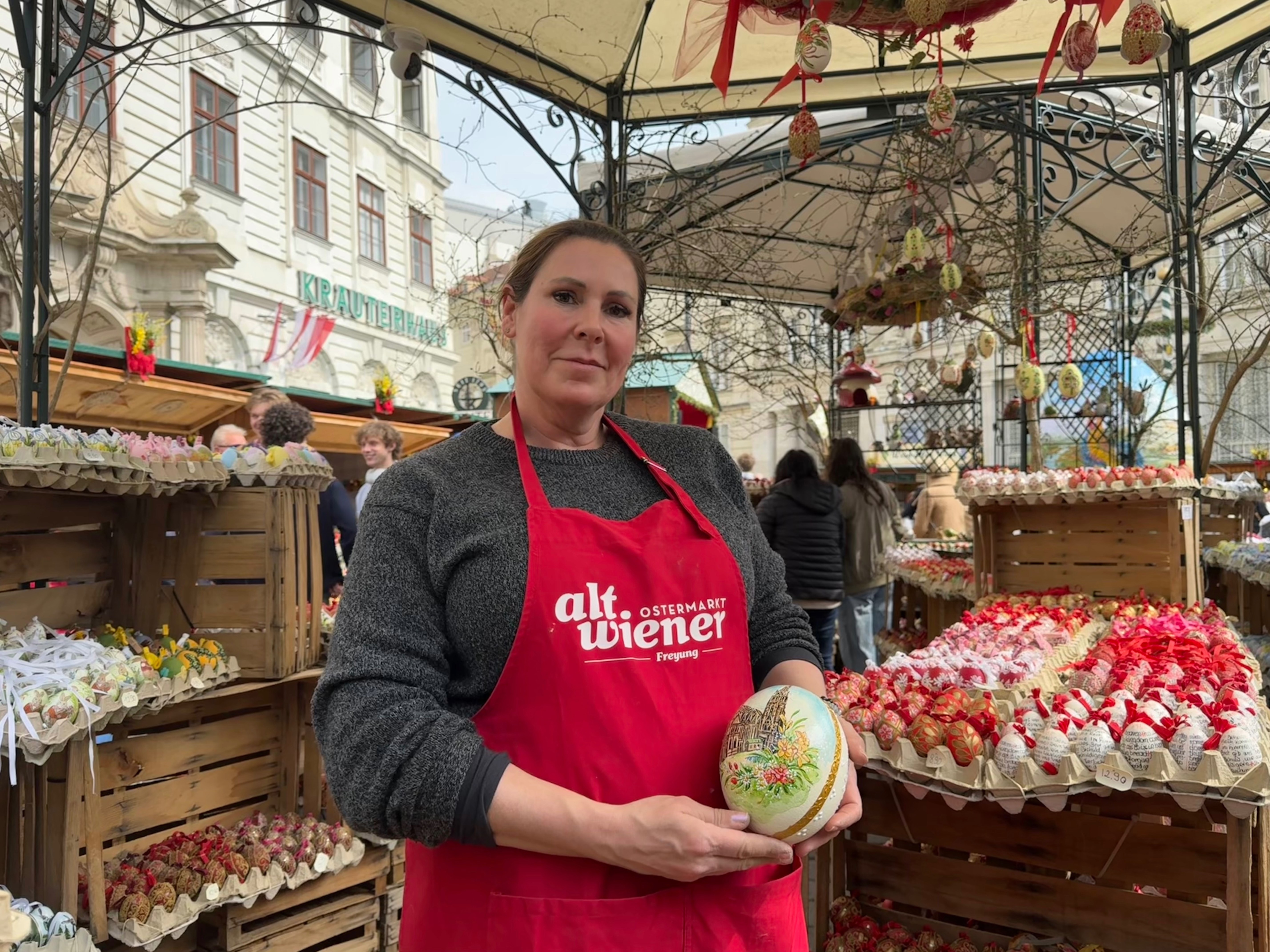 A woman in a red apron holds a decorated egg at an outdoor market stall filled with colorful ornamental eggs.