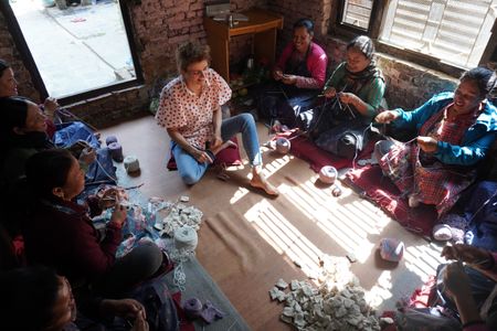 A group of women sitting on the floor in a sunlit room, engaged in a knitting activity with yarn balls scattered around.