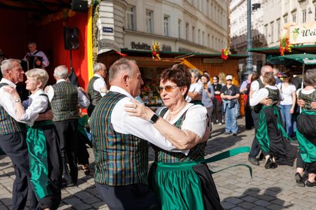 Older couples in traditional attire dance in a lively outdoor festival with wooden stalls and spectators in the background.