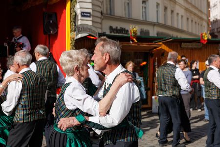 Elderly couples dance in traditional attire at an outdoor festival, surrounded by wooden stalls and a live band.