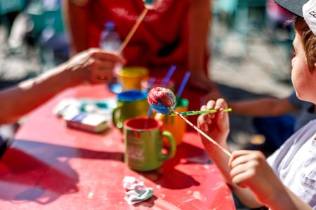 Children painting eggs with brushes at a red table, surrounded by colorful paint cups and sunlight.