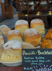 A display of Austrian Buchteln, soft golden-brown buns dusted with powdered sugar, sits on a plate. A sign describes them as plum jam-filled sweet donuts.