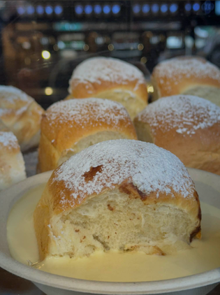 Fluffy rolls dusted with powdered sugar sit on a creamy sauce in a bowl. The background is softly blurred, evoking a cozy, inviting atmosphere