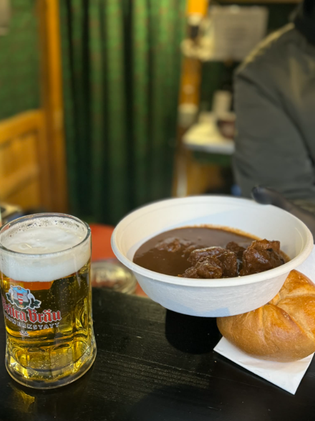 A bowl of rich brown stew with chunks of meat and a crusty roll next to a frothy glass of beer on a wooden table, evoking warmth and comfort.