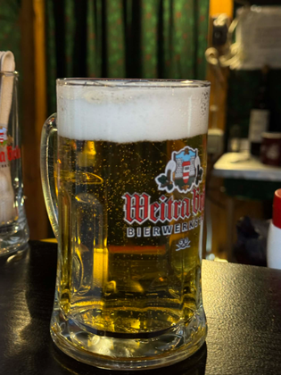 A glass mug filled with golden beer, topped with foam, stands on a wooden table. The label reads "Weitra Bier," and the background is softly blurred.