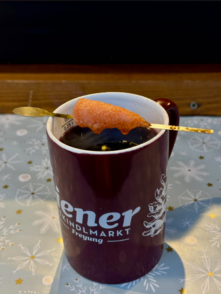 A festive maroon mug with white decorative text holds a warm drink. A sugar-coated pastry on a stick rests across the top, evoking a cozy winter vibe.