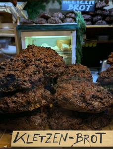 Stacks of dark, rustic Kletzenbrot loaves are displayed at a market stall, conveying a cozy, artisanal atmosphere. A sign in front reads "Kletzen-Brot."