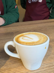 A creamy cappuccino in a white mug on a wooden table, with two people wearing red aprons in the background, creating a warm and inviting atmosphere.