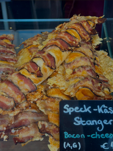 Crusty bread sticks wrapped with crispy bacon and melted cheese are stacked in a display case. A black sign reads "Speck-Käse Stangerl" with pricing details.