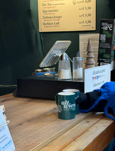 A rustic wooden counter with a green mug labeled "Alt Wien" alongside sugar containers. Menus and signs list hot drinks, liqueurs, and card payments accepted. Cozy, warm atmosphere.