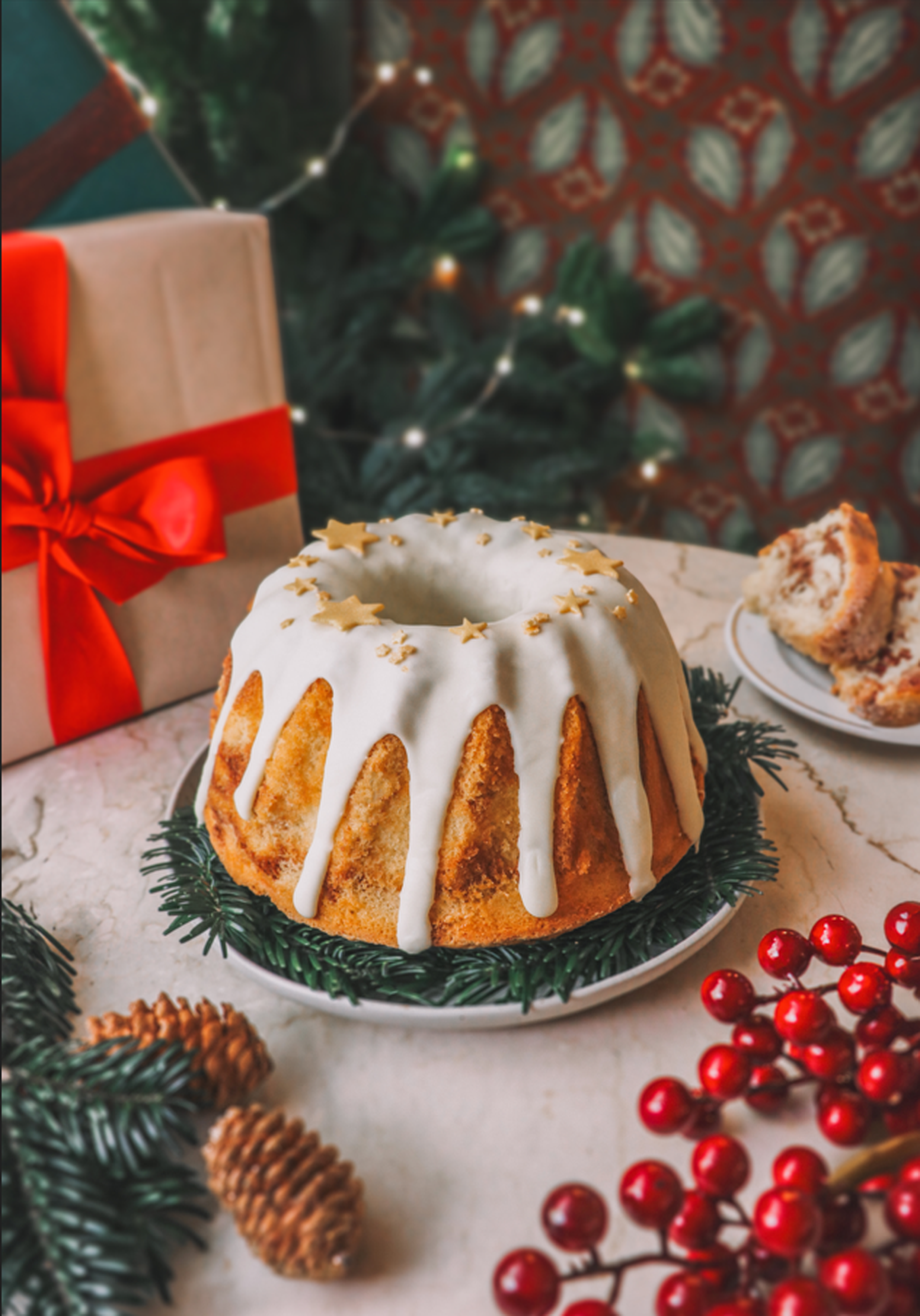 Festive bundt cake with white icing and star sprinkles, surrounded by holiday decorations, pinecones, and a gift box with a red ribbon.