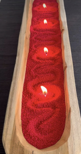 Long wooden tray filled with red sand and three lit candles evenly spaced, creating a warm and inviting atmosphere.