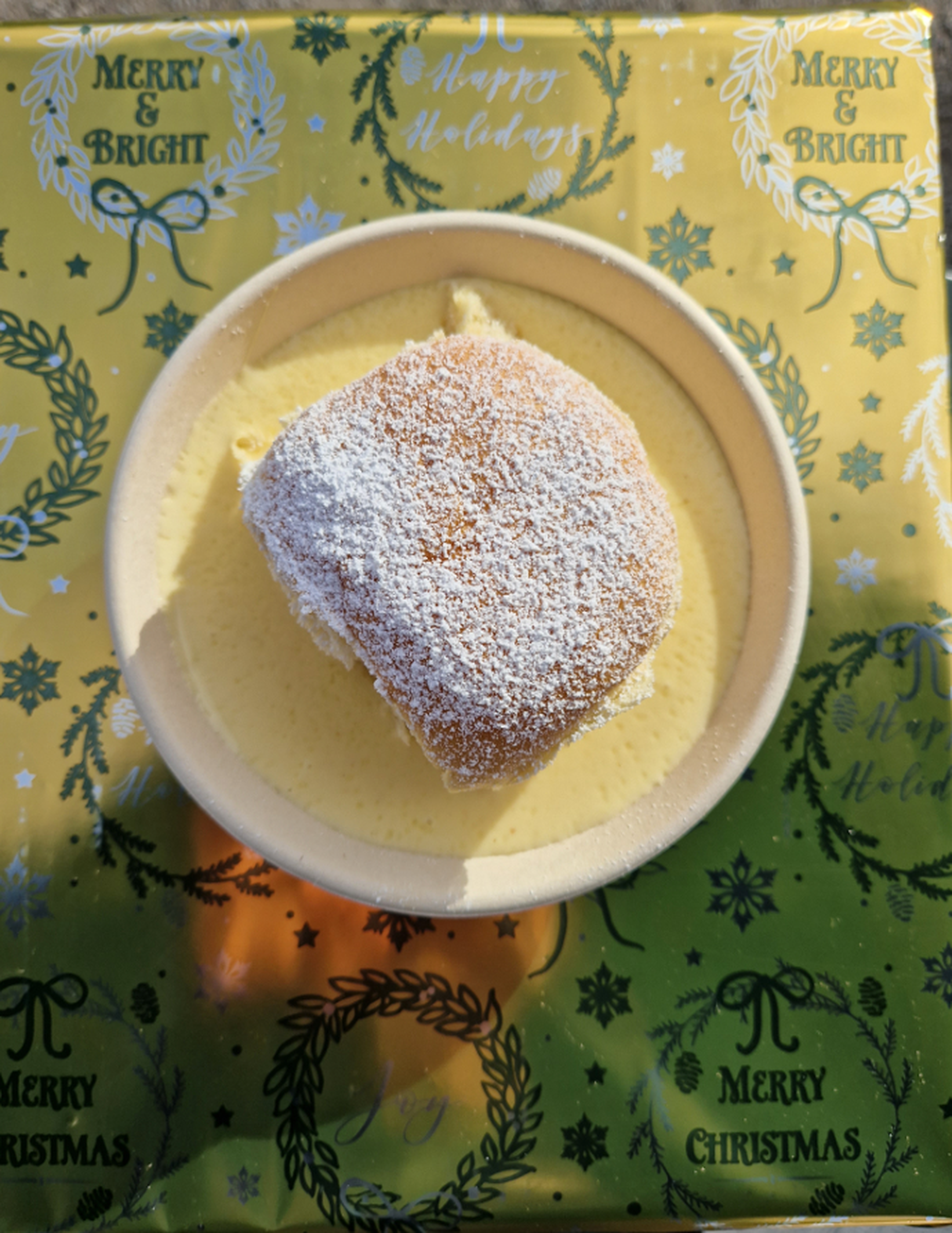 A powdered sugar-topped pastry sits on a plate of custard, placed on a festive "Merry Christmas" tablecloth.