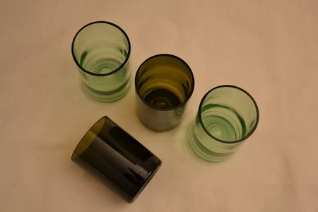 Four glass cups with various shades of green are arranged on a light beige surface, three upright and one laying on its side.