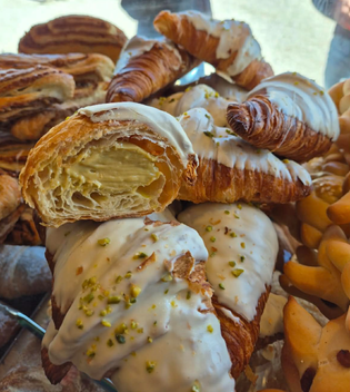 A display of croissants with white icing and crushed pistachios, revealing a creamy filling inside one. The scene conveys a tempting, bakery-fresh allure.