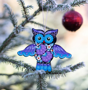 Colorful owl ornament with floral patterns hanging on a frosted pine branch, with a red bauble in the background.