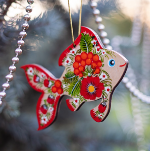 Wooden fish ornament with red floral patterns and a googly eye, hanging on a Christmas tree with silver bead garland.