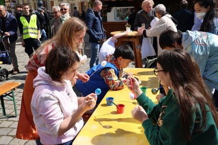 People at outdoor tables painting Easter eggs, with children and adults engaged in the activity. A lively, sunny atmosphere surrounds them.