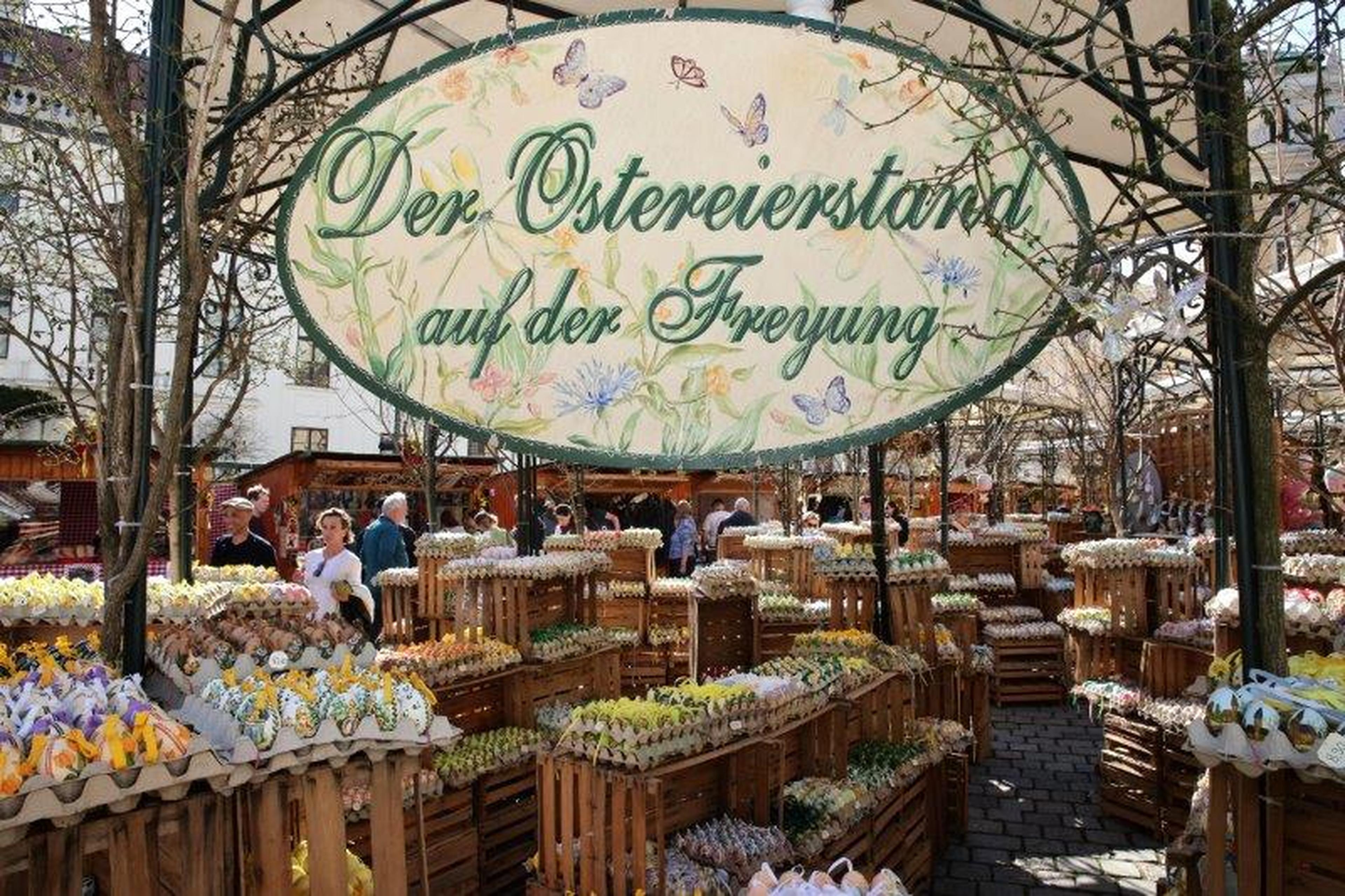 Bustling outdoor market with a large decorative sign, selling colorful eggs displayed on wooden stands under a canopy.