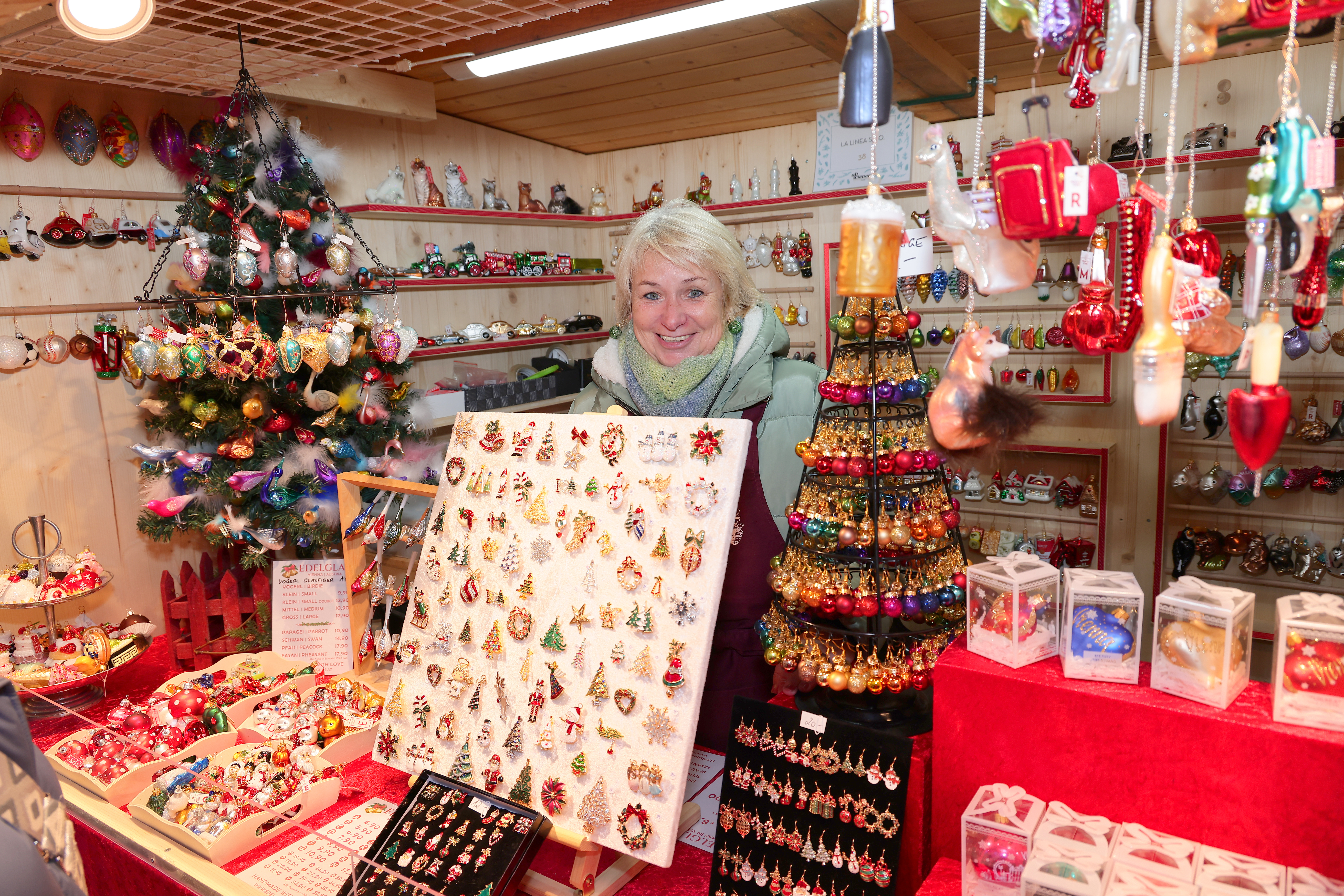 A woman at a festive market stall selling Christmas ornaments, with a decorated tree and colorful displays of baubles and trinkets.